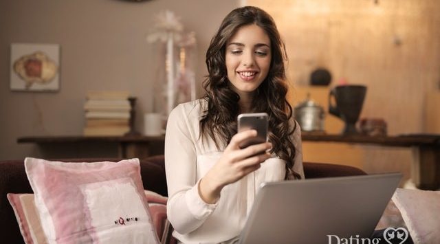 woman looking at phone while holding laptop